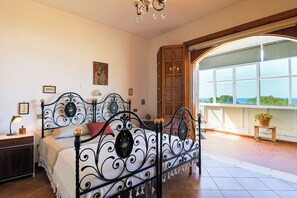 Ein Schlafzimmer mit einem schmiedeeisernen Bett, einem Nachttisch, einer Lampe und Meerblick durch ein großes Fenster.