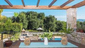 Een terras buiten met een houten pergola, een stenen zuil, rieten meubels, een witte vaas en uitzicht op een zwembad en groen.