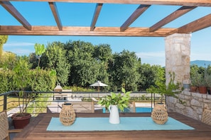 An outdoor patio with a wooden pergola, a stone pillar, wicker furniture, a white vase, and a view of a swimming pool and greenery.