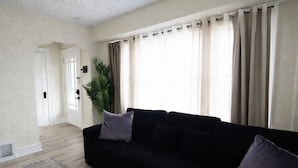 A living room with a black sofa, a grey rug, a potted plant, and beige curtains. - Modern Duplex Near Airport & Downtown + Game Room & Lake (Milwaukee)