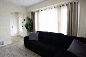 Ein Wohnzimmer mit einem schwarzen Sofa, einem grauen Teppich, einer Topfpflanze und beigen Vorhängen.