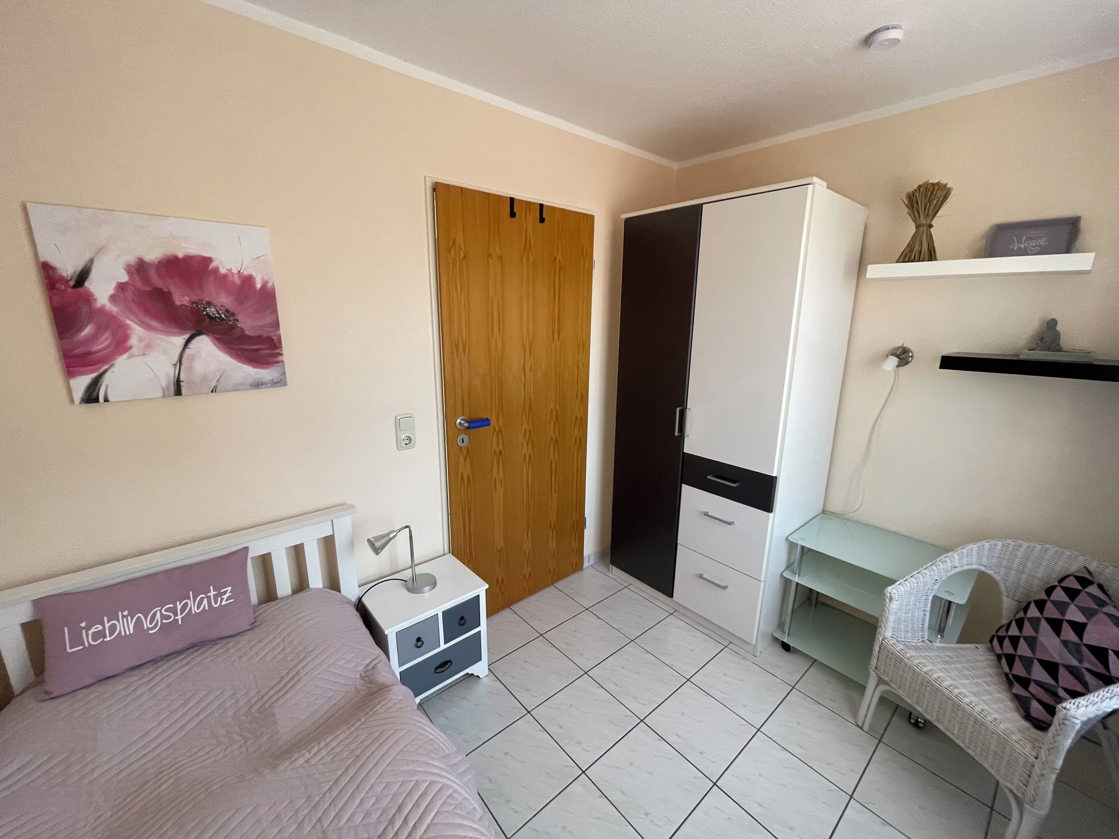 A bedroom with a bed, a wardrobe, a desk, a chair, a nightstand, and a wall-mounted shelf.