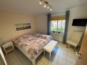 Ein Schlafzimmer mit Bett, Nachttisch, Bank, Fernseher und einem Fenster mit Vorhängen.