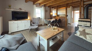 Un salón amplio con techo de madera, un televisor sobre un mueble, una mesa de comedor con sillas y una zona de cocina con estufa.