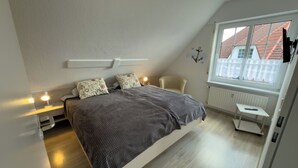 A bedroom with a bed, bedside lamps, a chair, a small table, and a window with a view of buildings. - Pelikan vacation apartment (Hooksiel)