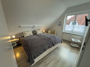 A bedroom with a bed, bedside lamps, a chair, a small table, and a window with a view of buildings. - Pelikan vacation apartment (Hooksiel)