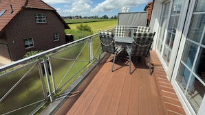 A balcony with a table and chairs, a view of a brick house, and a grassy field. - Pelikan vacation apartment (Hooksiel)