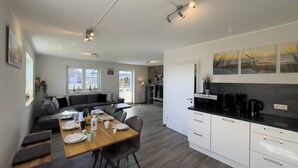 Une cuisine moderne avec des meubles blancs, un coin repas doté d’une table en bois et un salon avec un canapé de couleur foncée.