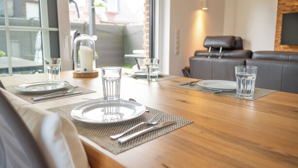 A dining table set with plates, cutlery, and glasses, with a view of a brick wall and a sofa in the background. - Stylishly furnished semi-detached house directly on the Wangermeer with garden, terrace, modern furnishings and dog-friendly - ideal for your North Sea vacation in Hohenkirchen. (Wangerland)