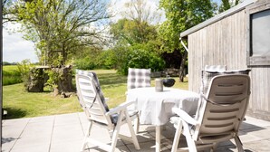 En udendørs terrasse med et bord og stole, omgivet af grønne planter og træer.