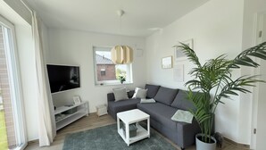 A modern living room with a grey sofa, a flat-screen TV, a white coffee table, and a large potted plant. - The "Genießerei" vacation apartment on the Wangermeer invites you to relax with its terrace, dog-friendly atmosphere and wallbox at the parking space. (Wangerland)