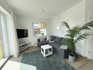 Ein modernes Wohnzimmer mit grauem Sofa, einem Flachbildfernseher, einem weißen Couchtisch und einer großen Küsterpflanze.