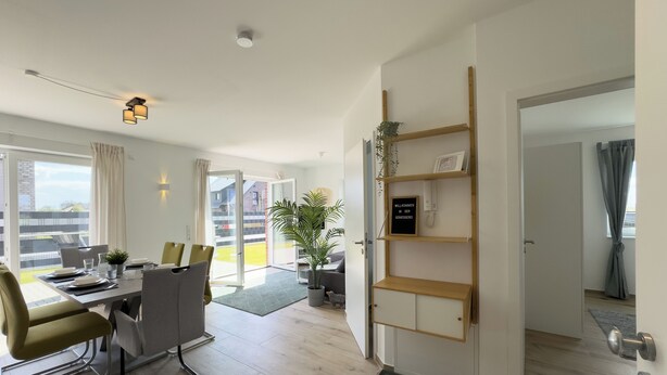 A modern living room with a dining area, a sliding glass door to a balcony, a wooden shelf unit, and a welcome sign. - The "Genießerei" vacation apartment on the Wangermeer invites you to relax with its terrace, dog-friendly atmosphere and wallbox at the parking space. (Wangerland)