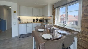 A modern kitchen with white cabinets, a dining area with a table set for four, and a view of buildings through a window. - Modern vacation apartment in Hooksiel with sunny balcony, two bedrooms, attic reading corner and quiet, central location near the North Sea. (Hooksiel)
