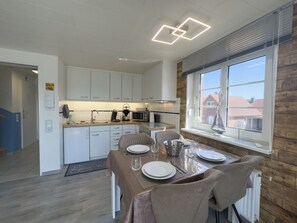 A modern kitchen with white cabinets, a dining area with a table set for four, and a view of buildings through a window. - Modern vacation apartment in Hooksiel with sunny balcony, two bedrooms, attic reading corner and quiet, central location near the North Sea. (Hooksiel)