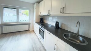 Moderni keittiö, jossa on valkoiset kaapit, musta työtaso, ruostumattomasta teräksestä valmistettu pesuallas ja ikkuna, josta avautuu näkymä taloon.