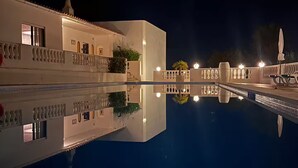 Vue nocturne d’un hôtel avec une piscine, un bâtiment blanc doté de balcons et un pont enjambant la piscine.
