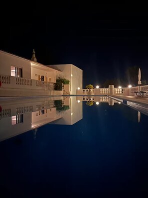 Ein Nachtbild eines Hotels mit Schwimmbad, ein weißes Gebäude mit Balkonen und eine Brücke über dem Pool.