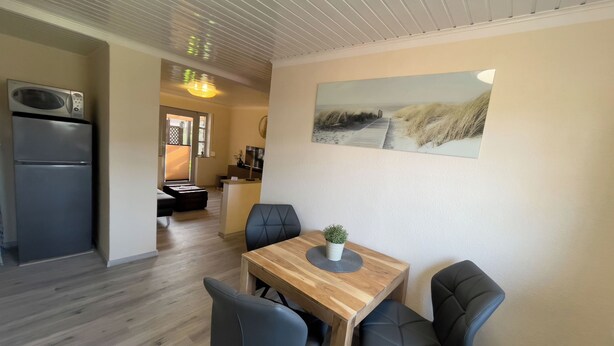A modern kitchen with a built-in microwave, a dining area with a wooden table and four chairs, and a painting of a beach scene on the wall. - Cozy vacation home with garden, terrace and central location - the "Kleine Hookie" in Hooksiel offers relaxation for couples, families and dog owners. (Hooksiel)