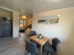 Une cuisine moderne équipée d’un four à micro-ondes encastré, un coin repas avec une table en bois et quatre chaises, et un tableau représentant une scène de plage accroché au mur.
