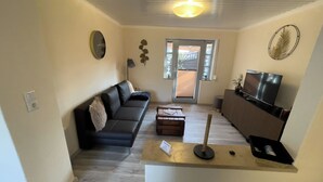 A living room with a sofa, a coffee table, a TV, and a small table with a lamp. - Cozy vacation home with garden, terrace and central location - the "Kleine Hookie" in Hooksiel offers relaxation for couples, families and dog owners. (Hooksiel)