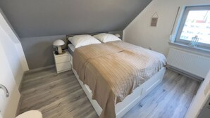A bedroom with a sloped ceiling, a bed with a beige bedspread, a nightstand with a lamp, and a window with a view of the outside. - Cozy vacation home with garden, terrace and central location - the "Kleine Hookie" in Hooksiel offers relaxation for couples, families and dog owners. (Hooksiel)
