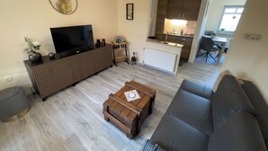 A living room with a sofa, a coffee table, a TV, and a kitchen in the background. - Cozy vacation home with garden, terrace and central location - the "Kleine Hookie" in Hooksiel offers relaxation for couples, families and dog owners. (Hooksiel)