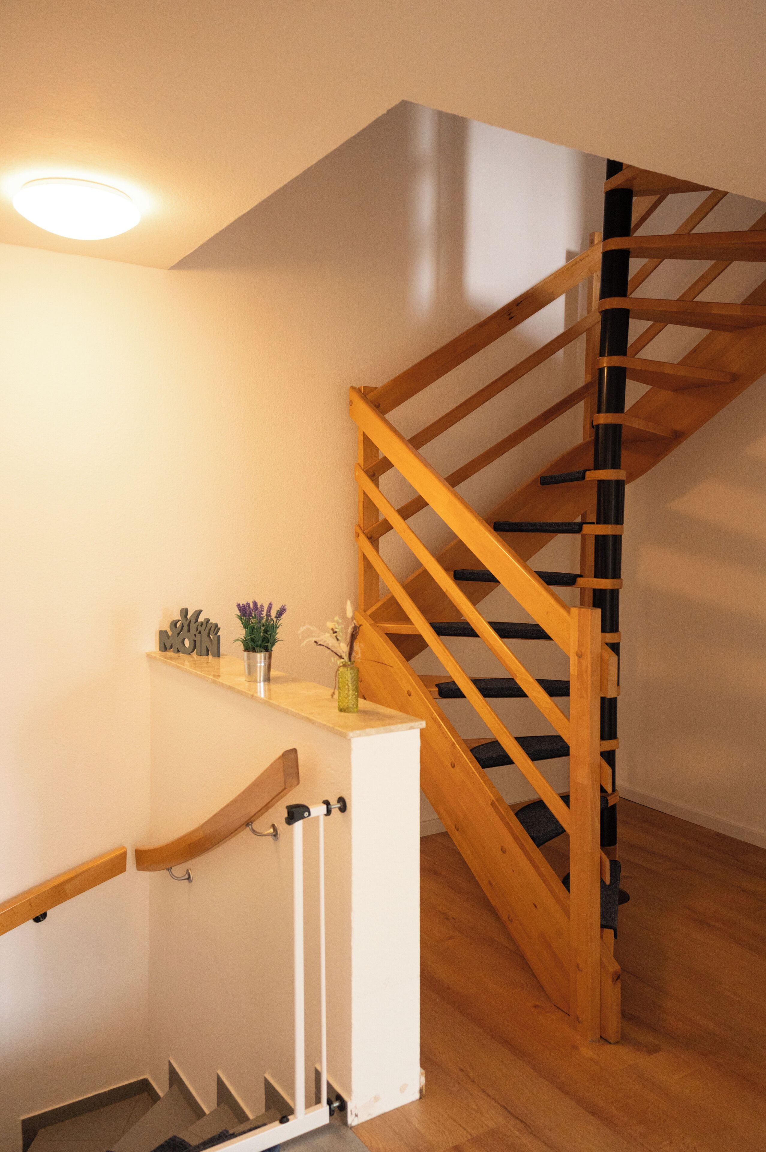 Un escalier en bois avec une rampe, une petite étagère ornée d’objets décoratifs et un luminaire fixé au mur.