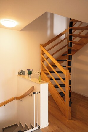 Un escalier en bois avec une rampe, une petite étagère ornée d’objets décoratifs et un luminaire fixé au mur.
