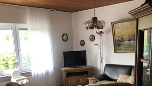 A living room with a television, a sofa, a dining table with chairs, a window with curtains, and a painting on the wall. - Bungalow close to the beach in Schillig with garden, three bedrooms, two bathrooms, up to three parking spaces and dog-friendly facilities - ideal for families and friends. (Schillig)