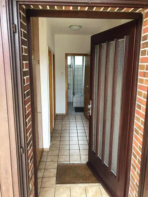 Käytävän lattia on laatoitettu, siinä on lasiruudullinen ovi, ja siitä on näkymä kylpyhuoneeseen, jossa on suihku ja laatoitetut seinät.