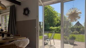 Et værelse med et spisebord, stole, en fletstol med hvid pude, en ophængt krans på væggen, og en skydedør af glas til en udendørs patio med græsplæne, stole og en dekorativ hængelygte.