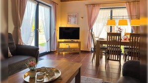 Een woonkamer met een bank, een bijzettafeltje met dienblad, een televisie op een standaard, een eettafel met stoelen en grote ramen met gordijnen.