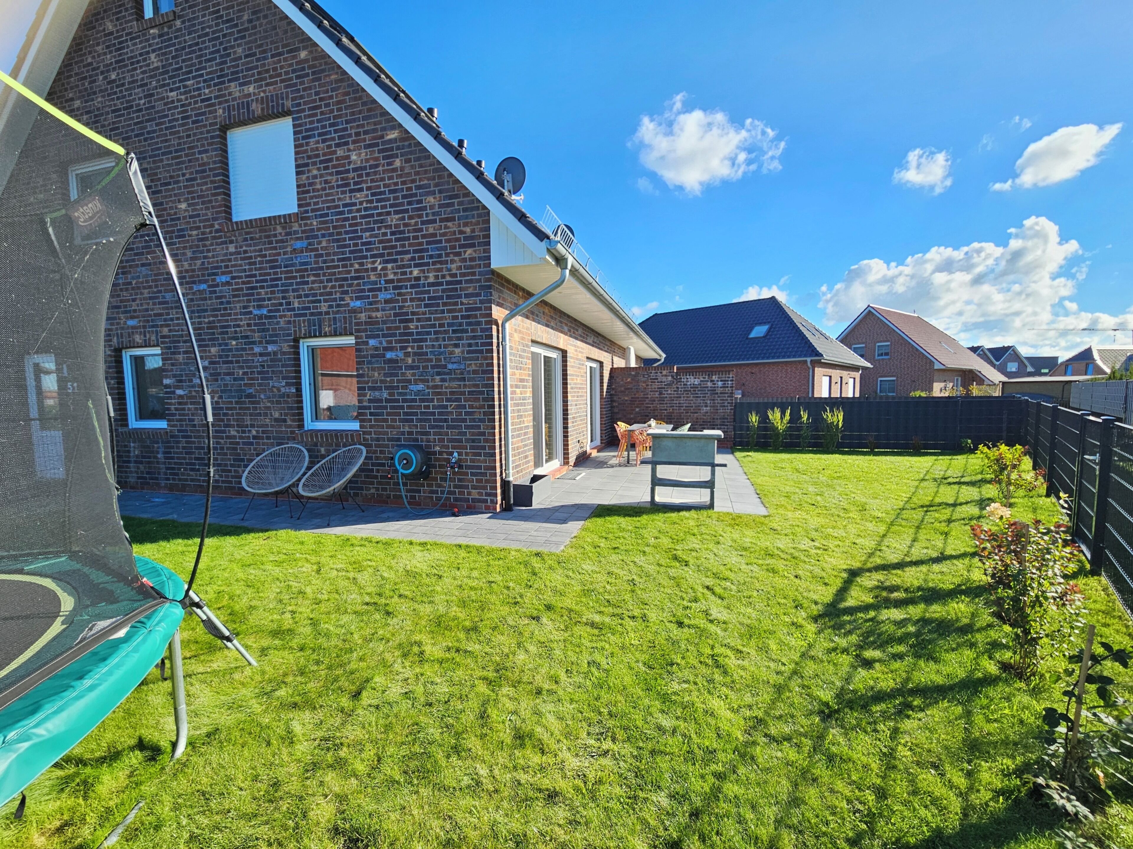 A residential backyard with a trampoline, a brick house, a patio with outdoor furniture, and a well-maintained lawn.
