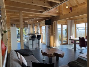 Ein geräumiger Raum mit Holzbalkendecke, einer Essecke mit Tisch und Stühlen sowie einer Küchenzeile mit Arbeitsfläche und Barhocker, und großen Fenstern mit Blick ins Freie.