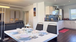 A modern dining area with a white table set for four, a kitchen in the background, and a cityscape view through a window. - Modern vacation home directly on the Wangermeer. Comfort and maritime flair for families, couples and dog owners. (Wangerland)