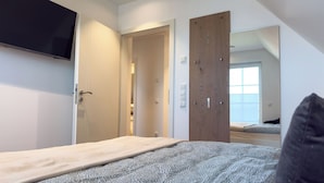 A bedroom with a bed, a TV mounted on the wall, and a wooden door. - Modern vacation home directly on the Wangermeer. Comfort and maritime flair for families, couples and dog owners. (Wangerland)