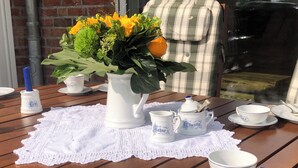 Een houten tafel gedekt met witte borden, kopjes en een vaas met bloemen, met een bakstenen muur en ramen op de achtergrond.