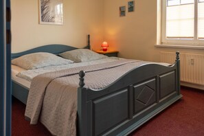 A bed with a headboard, a bedside table with a lamp, and a window with blinds. - Cozy vacation apartment in a quiet location in Hooksiel - ideal for families and couples looking for relaxation by the North Sea. (Hooksiel)