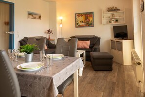 A dining area with a table set for two, a sofa, a TV, and a shelf with various items. - Cozy vacation apartment in a quiet location in Hooksiel - ideal for families and couples looking for relaxation by the North Sea. (Hooksiel)