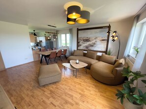 Ein modernes Wohnzimmer mit Holzboden, einem Ecksofa, einem Esstisch mit Stühlen und Pendelleuchten.