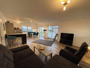 A modern living room with a sofa, a coffee table, a TV, and a dining area with a table and chairs. - Modern bungalow directly on the Wangermeer with fenced garden, wooden sauna, two bathrooms, fully equipped kitchen - ideal for families, couples and vacations with dogs. (Wangerland)
