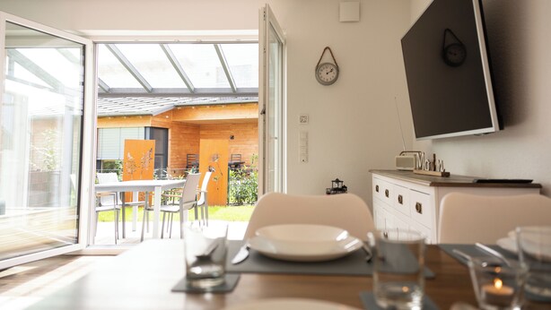 Et moderne spiseområde med et bord dækket til to personer, et fjernsyn monteret på væggen og en skydedør, der fører ud til en udendørs terrasse.