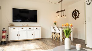 En moderne stue med et fladskærms-tv, hvide skabe, et spiseområde med stole, en vase med blomster, en dekorativt skibsror og en skulptur af et fyrtårn.