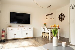 A modern living room with a flat-screen TV, white cabinetry, a dining area with chairs, a vase with flowers, a ship's wheel decoration, and a lighthouse sculpture. - Modern vacation home with garden on the Wangermeer - Mee(h)rzeit offers comfort, style and relaxation for the whole family, including four-legged friends. (Wangerland)