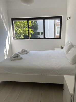 Un dormitorio con cama, ventana, mesita de noche pequeña y una planta en maceta.