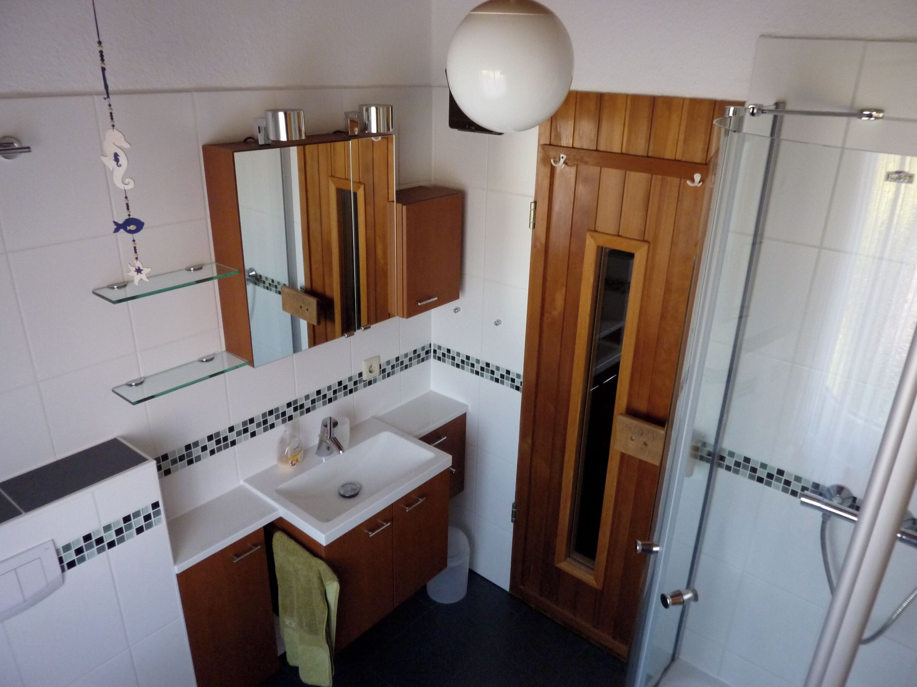 Een badkamer met een witte wasbak, houten wastafelmeubel, een spiegel, een douchecabine van glas en een hangende lamp.