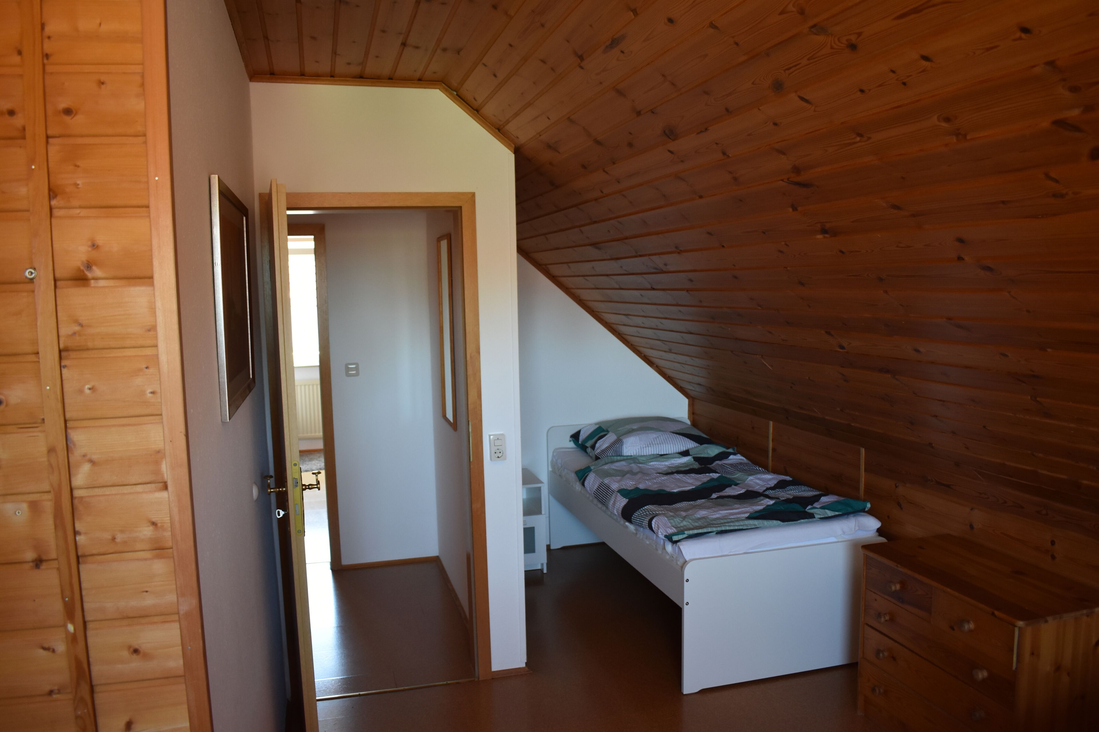 A small, single-bed room with a sloped ceiling, wooden walls, and a door leading to another room.
