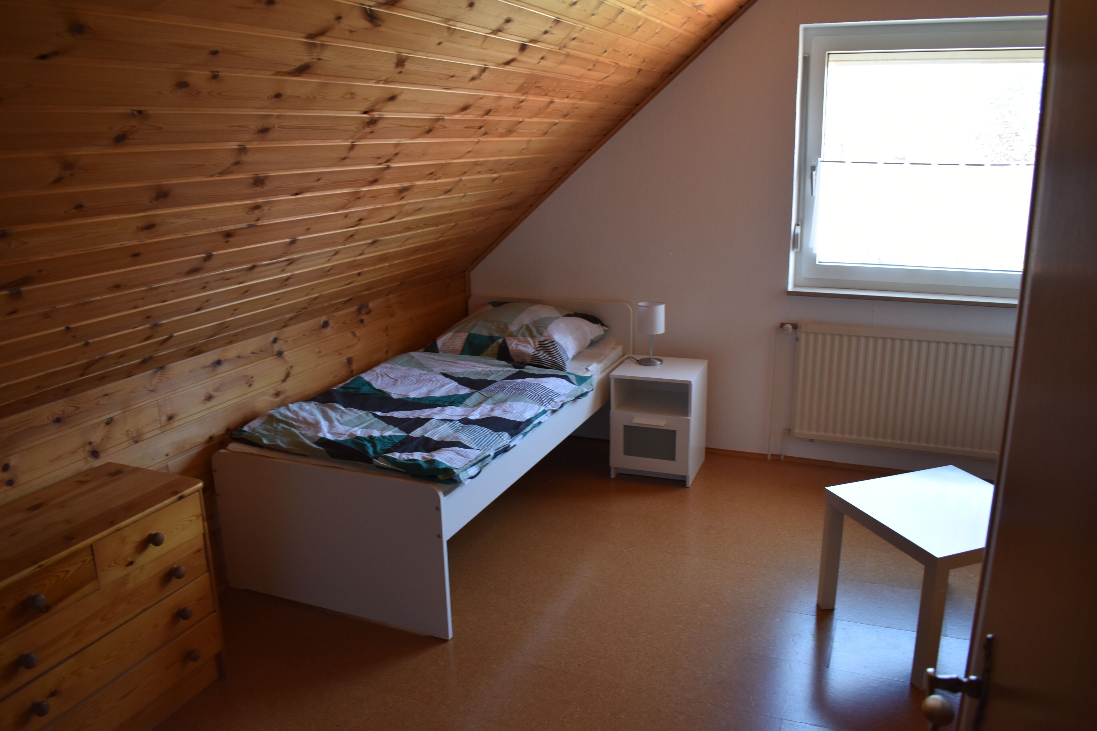 Ein Schlafzimmer mit schrägem Dach, einem Bett, einem Nachttisch, einem Heizkörper und einem Fenster.