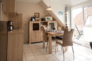 A modern kitchen with wooden cabinets, a dining table with chairs, and a large window with a view of a red-roofed building. - Stylish vacation apartment with balcony, modern furnishings and central location in Hooksiel - the Nordseeperle invites you to enjoy a relaxing vacation by the North Sea. (Hooksiel)